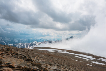 Awesome top view to high snowy mountain in dense low clouds. Scenic mountain landscape with beautiful peaks in thick clouds. Atmospheric view from stone hill to snow mountain tops among low clouds.
