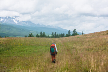 Traveling man in red with big backpack on way to snowy mountain range in low clouds. Backpacker walks through forest to large mountains under cloudy sky. Dramatic landscape with tourist in mountains.