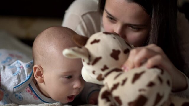 Newborn Active Baby With Young Mom. Cute Kid Smiling Teethless Face Portrait Early Days On Stomach Developing Neck Control. Mother And Child Look At Camera Play with Toy. Infant, Beginning Concept