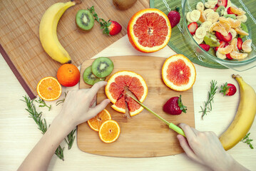 A child's hands are cutting grapefruit on a cutting board. Ingredients for making fruit salad. Top view. Flat lay.