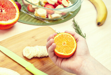 Child's hand holds juicy tangerine. Nearby are ingredients for making fruit salad. Blurred background.