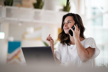 remote working dark haired woman calling and chatting infront of a laptop or notebook in casual outfit on her work desk in her modern airy bright living room home office with her mobile phone