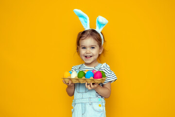 funny happy baby girl with easter eggs and bunny ears on yellow background