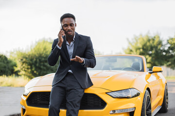 Portrait of rich African businessman wearing suit, standing near his luxury yellow cabriolet car...