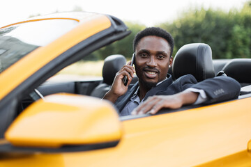 Handsome African American bearded business man in suit, sitting in his modern new car, talking on cellphone, smiling and looking at camera. Happy man owner is testing his new car