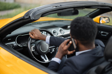 African Man in business suit using and talking on smartphone while driving a car, looking carefully at the road, back view.