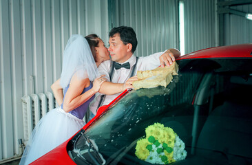 A young man with a bow tie and a woman in a veil are washing a car at a car wash. Wedding...