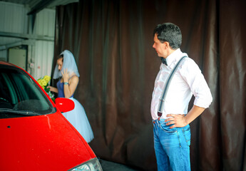 A young man with a bow tie and a woman in a veil are washing a car at a car wash. Wedding...