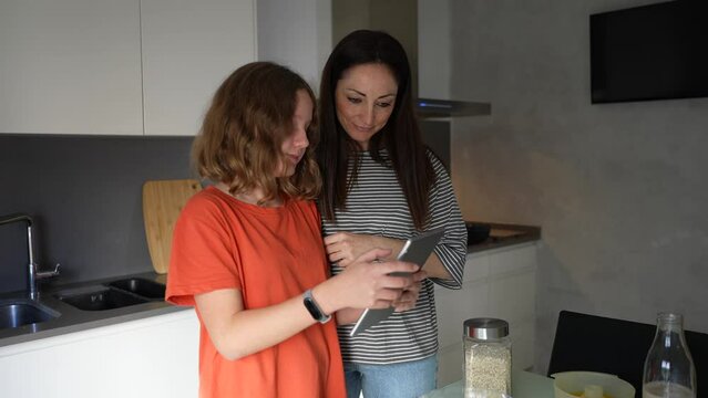 Happy mother and daughter preparing breakfast together at home, reading culinary blog on internet, using digital tablet, mother and teen child making healthy fruit drink