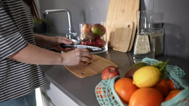 Close up of woman's hands cutting a lemon on a wooden cutting board preparing healthy breakfast, holding a knife and cutting fruits on the kitchen table.