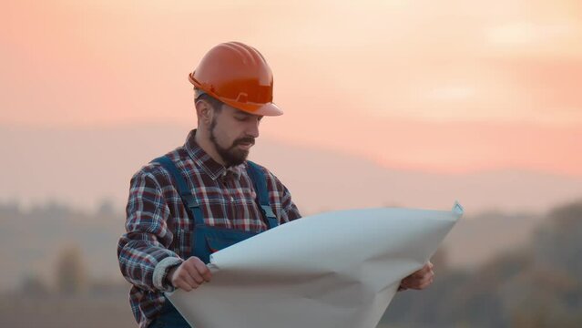 Close up builder architect reading draft in field. Man unrolling and examining blueprint while standing on field in countryside. At sunset. Slow motion.