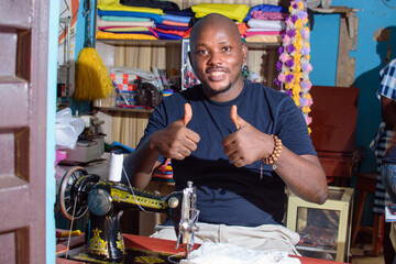 A happy African Nigerian male tailor, fashion designer or business man doing thumbs up gestures while making dress with a sewing machine in a tailoring shop