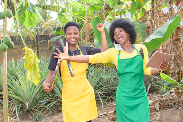 Two excited African female farmers or business women with colorful apron, holding farming tool and...