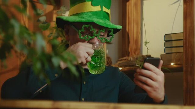 St Patrick day concept. Joyful man celebrating Saint Patricks day at home, having video call with friends. Man in hat and green glasses drinking green beverage using smartphone.