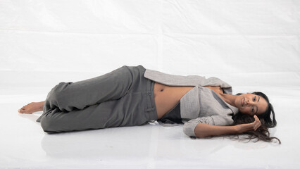 Tall glamorous woman in grey pants and jacket, reclining barefoot in a white studio setting.