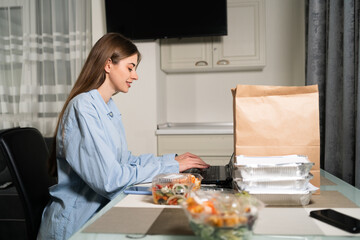 Obraz na płótnie Canvas paper bag and disposable takeaway food containers on the table, young woman ordering healthy food from laptop. Takeaway food, home delivery lunch