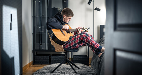 A young man plays the acoustic guitar in his room at home.