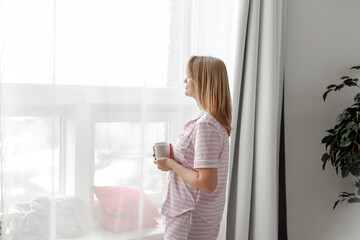 An attractive middle-aged woman in striped pink pajamas stands by the window with a cup of hot drink in her hands. Unhurried morning and slow life. Mental health and self-care. View from the back