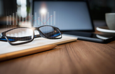 Work from home, desk office with digital tablet, blank notepad, eye glasses, on wooden table, close up and select focus on eye glasses with double exposure of investment chart