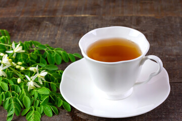 Moringa Tea in white ceramic cup with fresh green leaf and flower on wooden background. Moringa oleifera tropical herb healthy lifestyle concept.