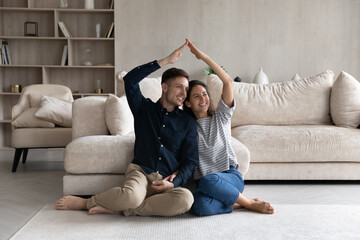 Happy young couple family making roof gesture, sitting on warm floor, smiling woman and man excited...