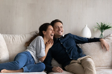 Excited dreamy man and woman hugging, sitting relaxing on couch at home, looking in distance, smiling girlfriend and boyfriend visualizing good future, spending lazy leisure time on sofa together