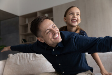 Obraz premium Smiling father piggy backing daughter, looking in distance, dreaming and having fun at home, happy loving young dad pretending plane with hands outstretched, family engaged in funny activity