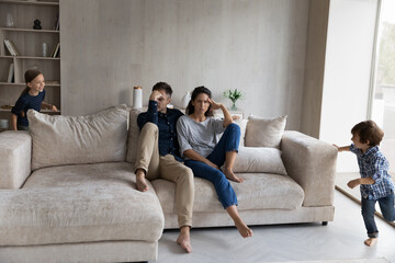 Tired parents sitting on couch at home while noisy little kids running and shouting around, young...