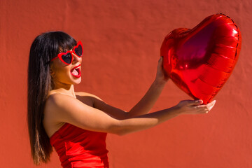 Brunette caucasian girl in love with heart balloons on Valentine's day in sunglasses, red background. The day of celebration a balloon