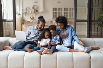 Happy friendly young African American couple parents and small adorable kids son daughter using different gadgets, playing games, web surfing, spending time online or shopping, modern tech addiction.