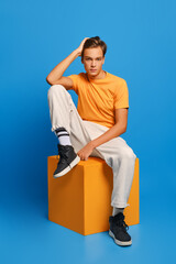 Positive man in orange undershirt and white sweatpants sits on wooden cube over blue background