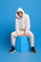 Young man in white sweatshirt with hood and white sweatpants sits on wooden cube over blue...