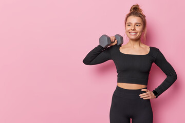 Sporty fit young woman has well developed muscles by strength training raises dumbbell has training in gym keeps hand on waist poses against pink background blank copy space for your promotion