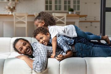 Joyful adorable laughing African American children son daughter lying on back of sincere smiling millennial father, playing together on big cozy sofa, enjoying family weekend playtime activity.