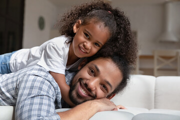 Happy cute small adopted kid daughter lying on back of sincere African american father, looking at camera resting on comfortable sofa together at home, single parenting, family relations concept.