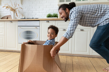 Happy adorable small curly kid boy sitting in carton box, looking at camera, playing entertaining pirates game with caring young African American father, having fun together in renovated kitchen.