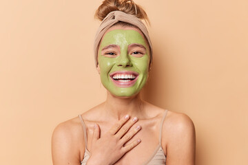 Happy European woman smiles broadly keeps hand on chest applies nourishing green mask undergoes beauty treatmentss wears headband has combed hair isolated over brown background. Skin care concept
