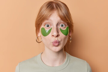 Portrait of surprised ginger girl keeps lips folded applies green collagen patches to reduce...
