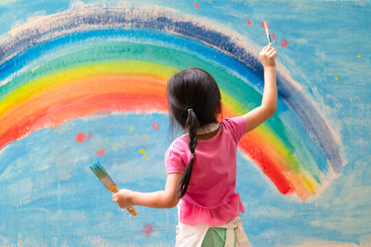 Unidentified little girl is painting the colorful rainbow and sky on the wall and she look happy and funny, concept of art education and learn through play activity for kid development.