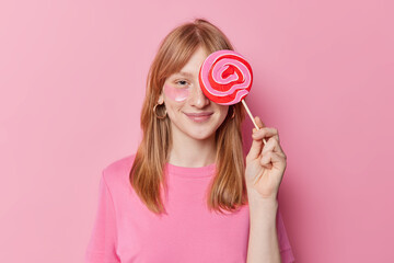Pleased redhead girl covers eye with big delicious candy applies beauty patches wears casual t shirt poses against pink background has sweet tooth. Children confectionary and beauty concept.