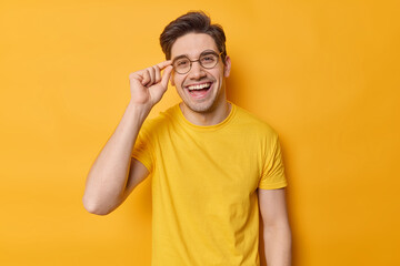 Waist up shot of cheerful brunet guy keeps hand on rim of spectacles wears casual t shirt and round...