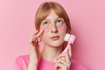 Pensive freckled millennial girl makes face massage with brush applies hydrogel patches under eyes...