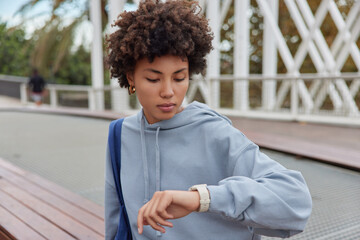 Outdoor shot of curly haired woman wears casual hoodie checks time on smartwatch carries bag waits for someone has serious expression waits for trainer to start sport training. People and lifestyle