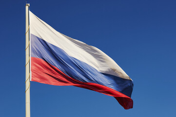 The national flag of the Russian Federation flutters in the wind against a blue cloudless sky. The tricolor symbol of the state, attached to the pole with a cord. Bright sunny day. Light and shadows.