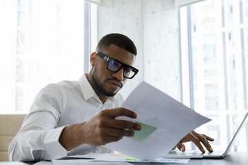 Serious African American manager in stylish glasses reading corporate document with note, sales...
