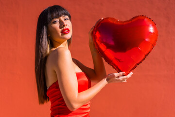 Brunette caucasian girl in love with heart balloons on Valentine's day, red background. Portrait on the day of celebration