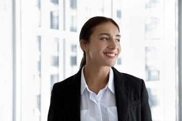 Happy young female business professional posing at office window glass with city view, looking away, smiling, thinking of future career growth, job success. Confident businesswoman head shot portrait