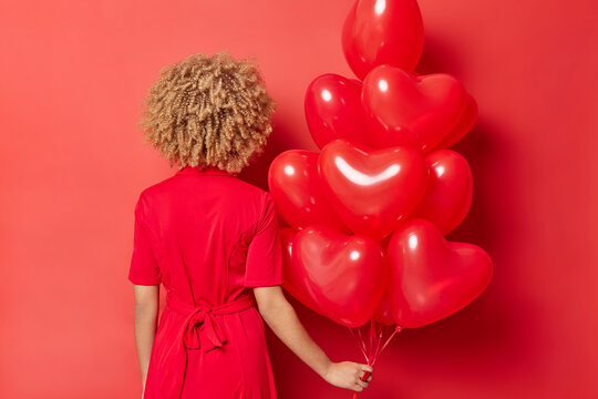 Faceless woman stands back to camera wears dress holds bunch of inflated heart shaped balloons for Valentines Day celebrates lovers holiday prepares for romantic party with boyfriend. Red background