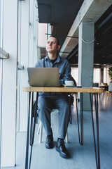 A young successful businessman in a suit drinks coffee and works in a cafe on wireless free internet on a laptop, sitting alone at a table in a cafe.