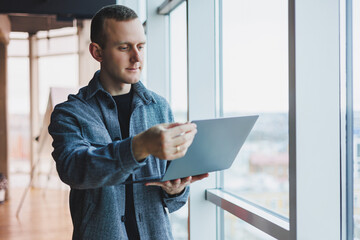 Portrait of a smart intelligent male manager, he is holding a laptop for research in the office, a business man in elegant clothes playing on the touchpad. Standing in the office by a large window